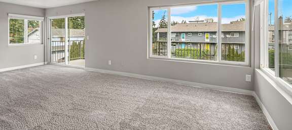 A spacious room with grey carpeting and large windows overlooking a residential area.